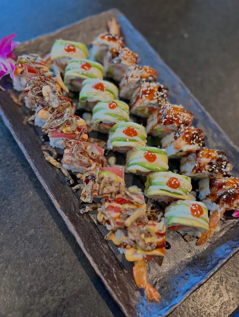 Three rows of specialty sushi rolls on a dark slate platter: tuna rolls with crispy onions, avocado-wrapped rolls, and eel-glazed rolls with sesame